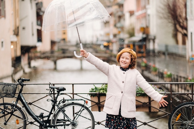 Hermosa chica alegre con un abrigo con un paraguas transparente en Annecy. Francia. La niña levanta alegremente un paraguas bajo la lluvia.