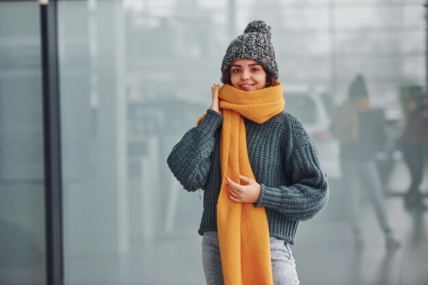 Hermosa chica alegre con bufanda amarilla y ropa de abrigo parada en el interior contra el fondo con reflejos.