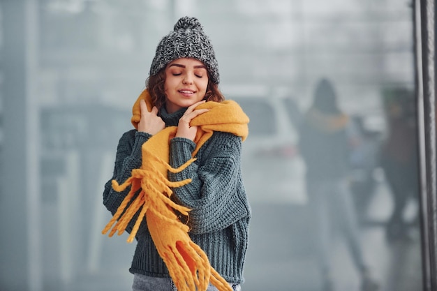 Hermosa chica alegre con bufanda amarilla y ropa de abrigo parada en el interior contra el fondo con reflejos.