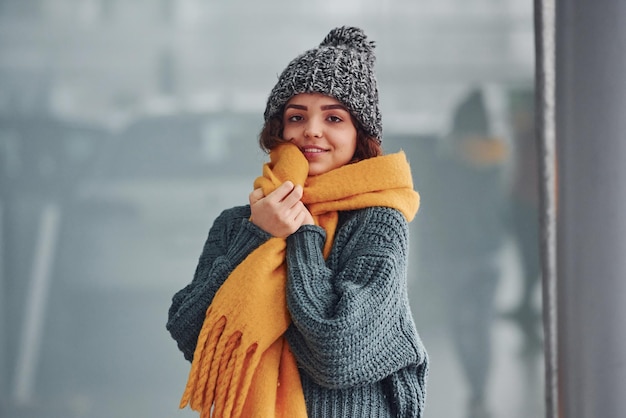 Hermosa chica alegre con bufanda amarilla y ropa de abrigo parada en el interior contra el fondo con reflejos.