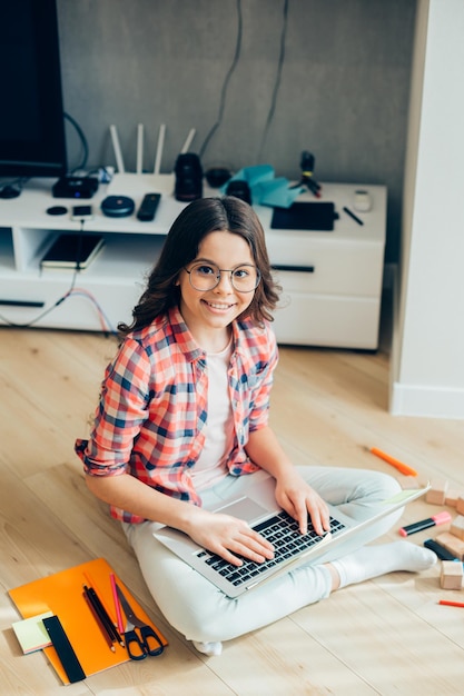 Hermosa chica alegre con gafas sentada en el suelo y sonriendo mientras sostiene una computadora portátil Suministros de papelería y ladrillos de madera a su lado