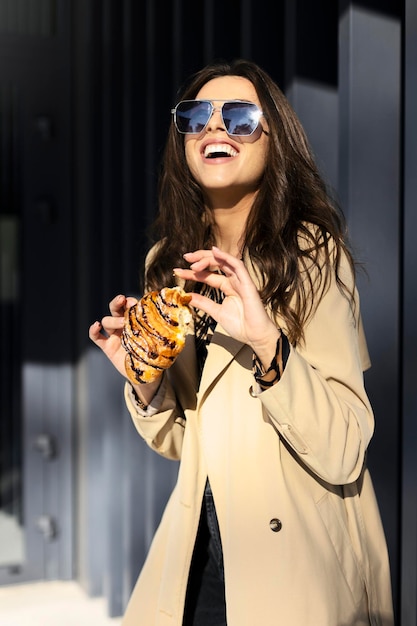 Hermosa chica alegre con gafas de sol come un croissant rompe un pequeño trozo Una joven en la calle sonriendo sinceramente levantó la cara hacia el sol