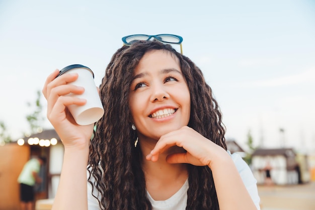 Una hermosa chica alegre con rastas está sentada en un patio de comidas y bebiendo café café de comida rápida en la calle una mujer bebe café en un café otoño