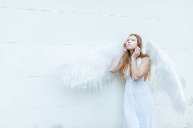 Hermosa chica ángel con alas blancas cerca de la pared mirando pensativamente hacia arriba