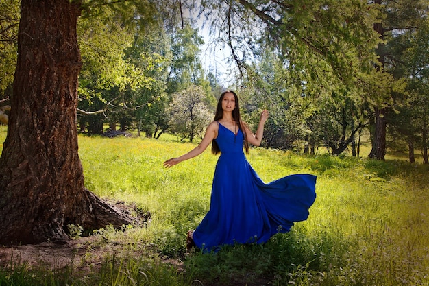 Una hermosa chica de apariencia asiática con un vestido azul baila bajo un árbol en las montañas de Altai. Concepto de turismo.