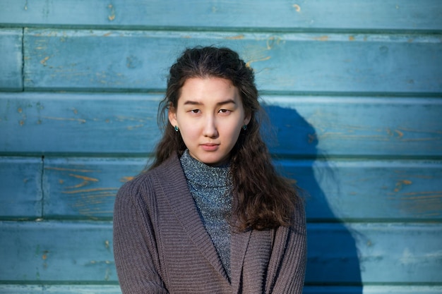 Una hermosa chica asiática con cabello largo y un suéter cálido está de pie junto a la pared azul de madera. Ella mira hacia adelante misteriosamente.