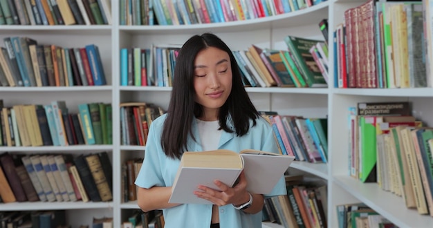 Hermosa chica asiática con camisa azul camina por los estantes con libros en la biblioteca Concepto de educación universitaria