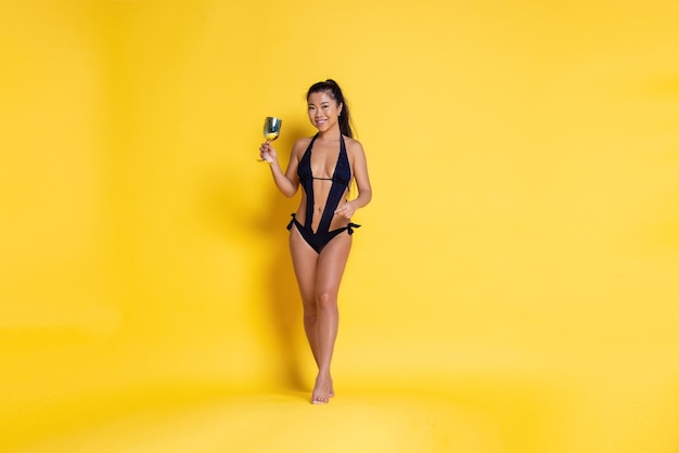 Hermosa chica asiática con copa de vino en la mano vistiendo traje de baño negro sobre un fondo amarillo