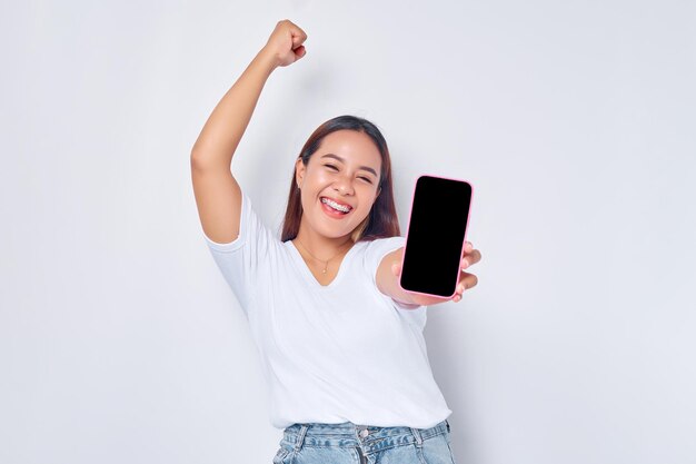 Hermosa chica asiática emocionada con una camiseta blanca casual que muestra un teléfono móvil con una pantalla blanca en blanco levantando el puño celebrando la buena suerte aislada en el concepto de estilo de vida de la gente de fondo blanco