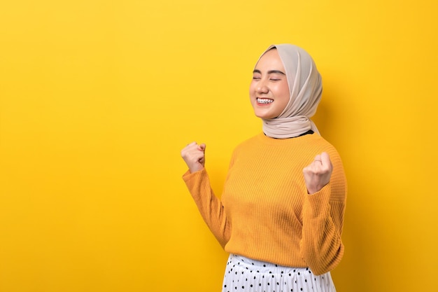 Hermosa chica asiática emocionada usando hiyab levantando el puño celebrando el éxito aislado en un fondo amarillo