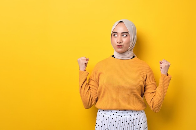 Hermosa chica asiática emocionada usando hiyab levantando el puño celebrando el éxito aislado en un fondo amarillo