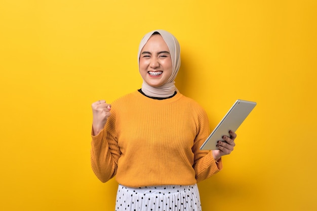 Hermosa chica asiática emocionada usando hiyab usando una tableta digital levantando el puño celebrando el éxito aislado en un fondo amarillo