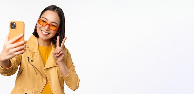 Hermosa chica asiática con estilo en gafas de sol tomando selfie en el teléfono inteligente posando para la foto con mobi