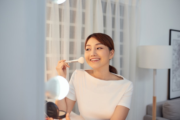 Hermosa chica asiática mirando en el espejo y aplicando cosmético con un pincel grande