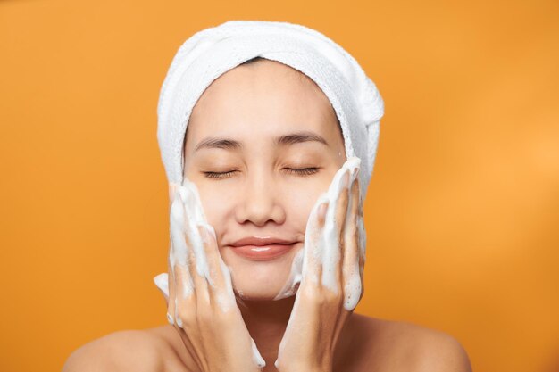 Hermosa chica asiática poniéndose crema en la cara aislada de fondo naranja