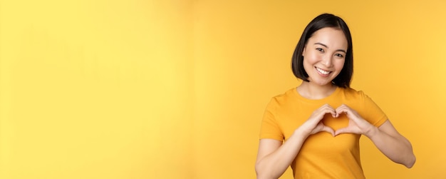 Hermosa chica asiática que muestra un gesto de amor al corazón y dientes blancos sonrientes expresa cuidado y simpatía de pie sobre un fondo amarillo