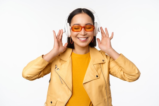 Hermosa chica asiática riendo feliz escuchando música en auriculares de pie sobre fondo blanco.