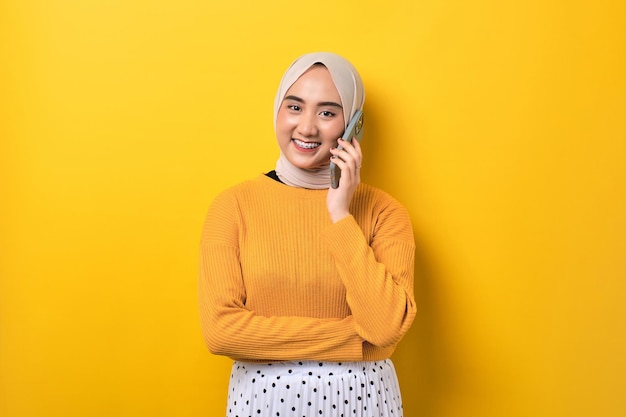 Hermosa chica asiática sonriente con hiyab hablando por teléfono móvil mirando a la cámara con expresión facial feliz aislada de fondo amarillo