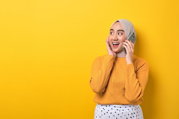 Hermosa chica asiática sorprendida usando hiyab hablando por teléfono celular con un amigo mirando el espacio vacío aislado en el fondo amarillo