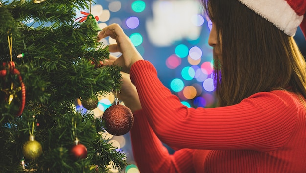 Hermosa chica asiática en suéter de santa que cuelga adornos coloridos y encantadores y una bola en el árbol de Navidad para decorar la hermosa fiesta de celebración en la noche feliz. Agregue un poco de ruido para adaptarse a la imagen de estilo vintage.