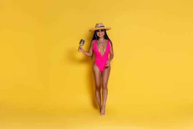 Hermosa chica asiática en traje de baño rosa y un sombrero con copa de vino sobre un fondo amarillo