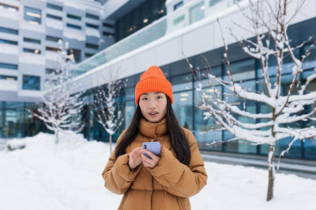 Hermosa chica asiática usa el teléfono para citas en línea hace una elección camina en un día nevado de invierno