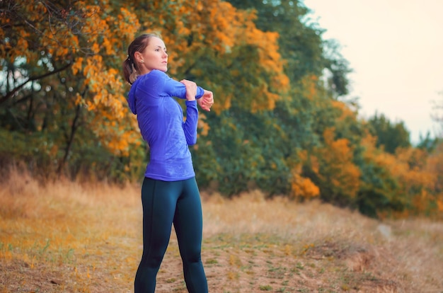 Hermosa chica atlética caucásica en camisa azul y leggins deportivos negros realiza calentamiento antes de trotar