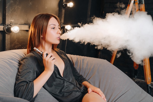 Hermosa chica atractiva exhalando vapor mientras fuma un cigarrillo electrónico, pasando el tiempo libre mientras descansa sobre un puf suave sentado en la sala de diseño.