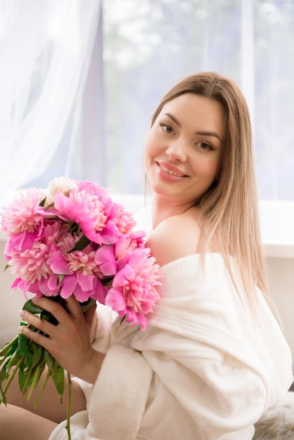 Hermosa chica en una bata blanca con un ramo de peonías rosas cerca del baño