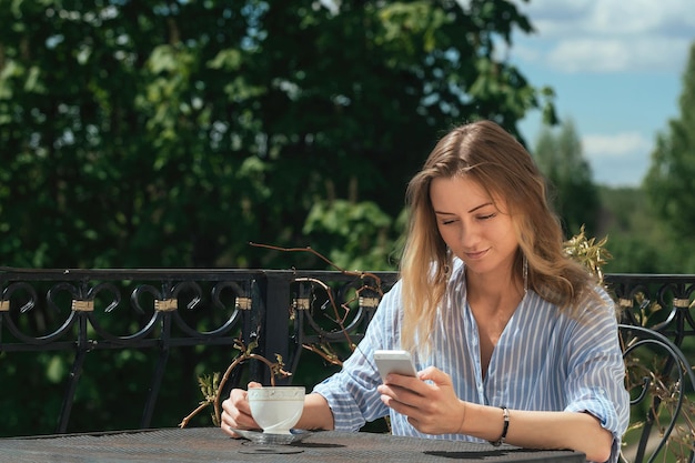 Hermosa chica bebe café en una mesa en el balcón de su casa y mira a través de su teléfono