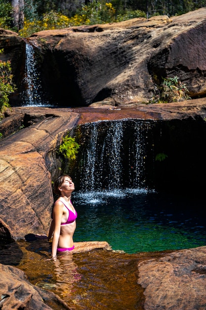 hermosa chica en bikini rosa se sienta junto a la piscina de roca natural con cascadas en la meseta negra