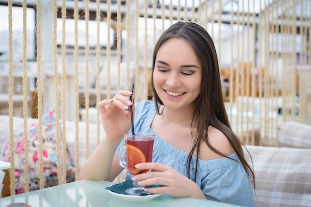 Hermosa chica en una blusa azul en un café con una copa de vino caliente riéndose de una mesa en un café