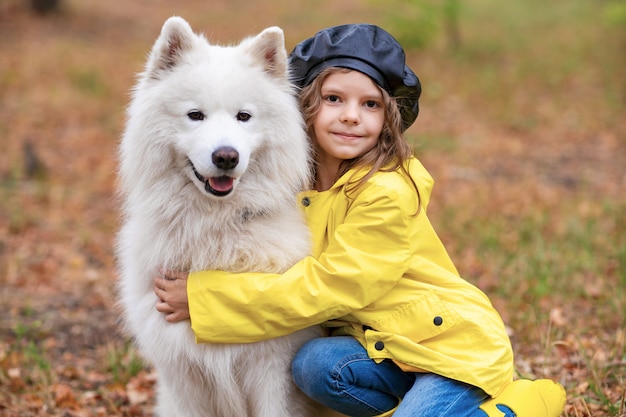 Hermosa chica con botas de goma amarillas y chubasquero en un paseo, juega con un hermoso perro samoyedo blanco en el parque de otoño