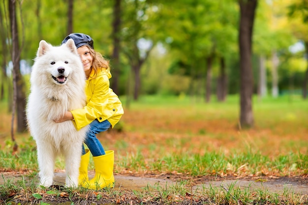 Hermosa chica con botas de goma amarillas y impermeable en paseos, juega con un hermoso perro samoyedo blanco en el parque de otoño.