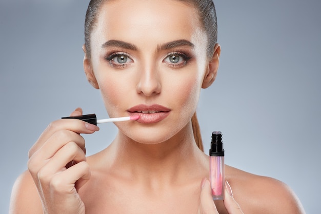 Hermosa chica con cabello castaño arreglado detrás, piel limpia y fresca, ojos grandes y hombros desnudos posando en el fondo gris del estudio, foto de belleza, usando brillo de labios, mirando a cámara.