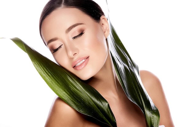 Hermosa chica con cabello castaño arreglado detrás, piel limpia y fresca, ojos grandes y hombros desnudos posando con hojas verdes en la pared blanca, de cerca.