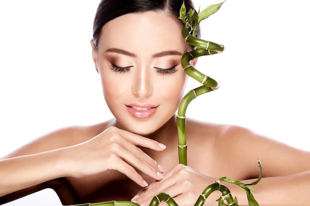 Hermosa chica con cabello castaño arreglado detrás, piel limpia y fresca, ojos grandes y hombros desnudos posando con planta verde en pared blanca, retrato, concepto de belleza.