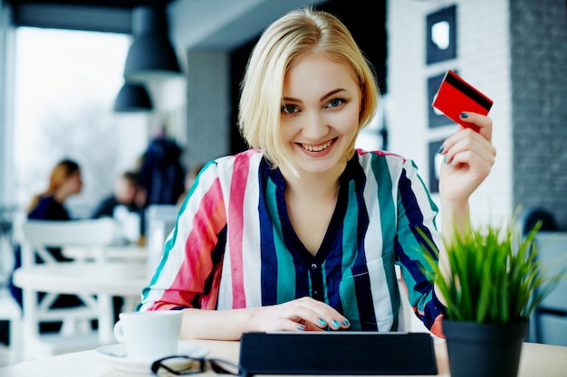 Hermosa chica con cabello claro con camisa colorida sentada en la cafetería con tableta, tarjeta de crédito y taza de café, concepto independiente, compras en línea.