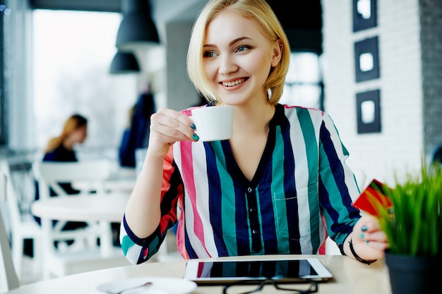 Hermosa chica con cabello claro con camisa colorida sentada en la cafetería con tableta, teléfono móvil y taza de café, concepto independiente, compras en línea.
