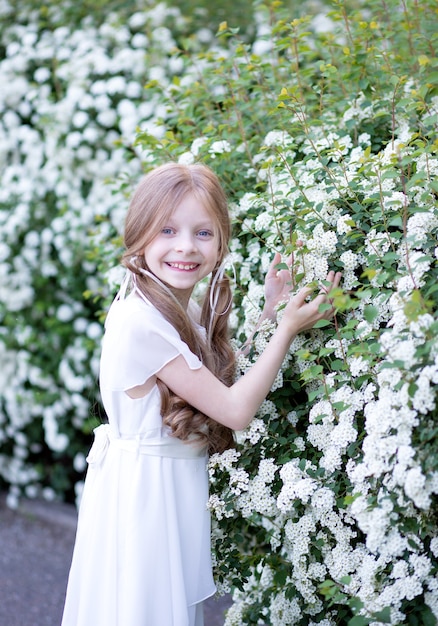 Hermosa chica con cabello largo rubio con un delicado vestido de seda blanco, contra las flores blancas de primavera, sostiene una flor, sonríe dulcemente y mira hacia el marco