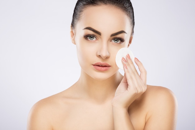 Hermosa chica con cabello negro arreglado detrás, ojos grandes, cejas gruesas y hombros desnudos de pie en el fondo gris del estudio y sosteniendo una esponja de limpieza cosmética, espacio de copia.