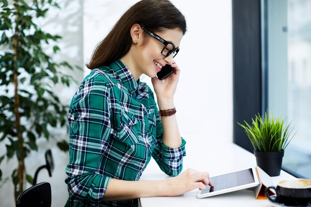 Hermosa chica con cabello negro con camisa y anteojos sentado en la cafetería con tableta y taza de café, sosteniendo el teléfono móvil, concepto independiente, sonriendo.