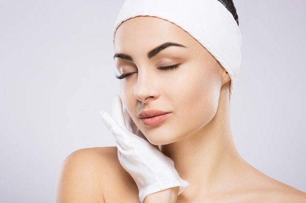 Hermosa chica con cabello negro fijo detrás, ojos grandes, cejas gruesas y hombros desnudos con vendaje para el cabello y guantes en la pared gris, foto de belleza, retrato.