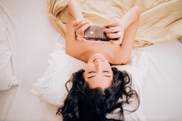 Hermosa chica con cabello ondulado negro desnuda tomando un selfie por teléfono acostado en la cama por la mañana. La toma interior de una alegre mujer caucásica tiene una videollamada matutina, tiene un teléfono celular moderno.