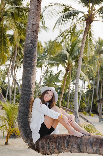 Hermosa chica de cabello oscuro feliz europea cerca de la palma de coco en la playa de arena blanca