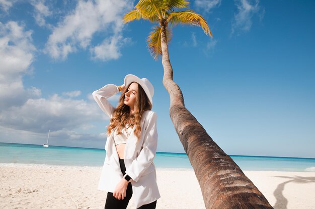 Hermosa chica de cabello oscuro feliz europea cerca de la palma de coco en la playa de arena blanca