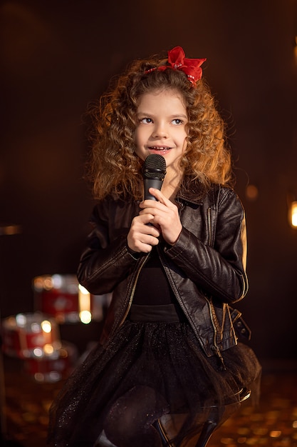 Hermosa chica con cabello rizado con chaqueta de cuero y gafas de sol rojas canta en un micrófono inalámbrico para karaoke en estudio de grabación
