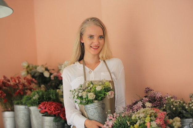 Hermosa chica de cabello rubio de pie con confianza frente a flor en tienda de flora de minoristas abiertos.