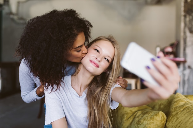 Hermosa chica con cabello rubio tomando fotos soñadoramente en el teléfono celular mientras una chica bastante afroamericana la besa en la mejilla mientras pasan tiempo juntos en casa