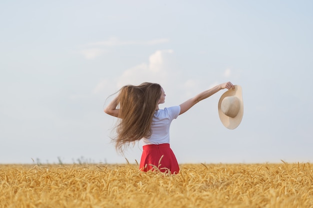 Hermosa chica camina en el campo y disfrutando día soleado. La mujer joven está sosteniendo el sombrero contra la pared del campo de trigo